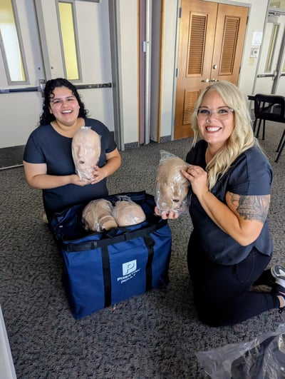 Two CPR instructors smile while unpacking training manikins from a blue Prestan training bag during a First Aid and CPR class setup. The scene is indoors, with classroom doors and chairs in the background. Two CPR instructors smile while unpacking training manikins from a blue Prestan training bag during a First Aid and CPR class setup. The scene is indoors, with classroom doors and chairs in the background.