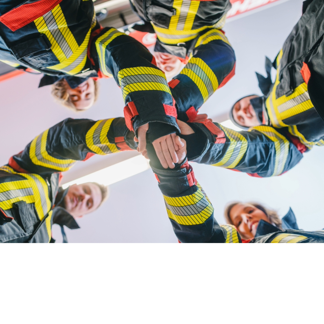 Group of firefighters in uniform standing in a circle with hands joined in the center, symbolizing teamwork and emergency readiness—ideal image for promoting Basic Life Support (BLS) training.