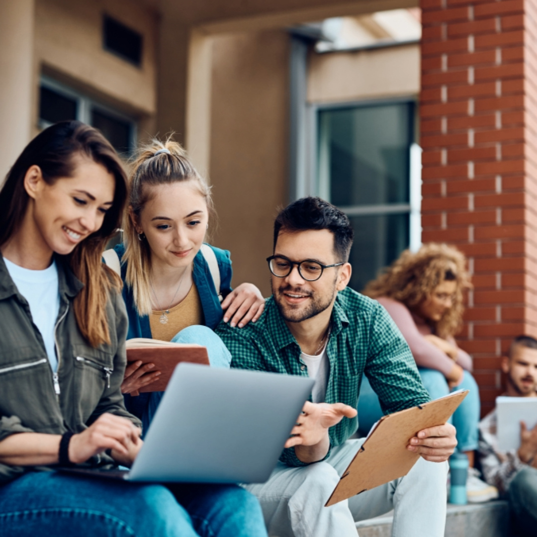 Group of diverse college students sitting on outdoor steps, smiling and using tablets and phones. Promoting First Aid and CPR training for educational organizations.